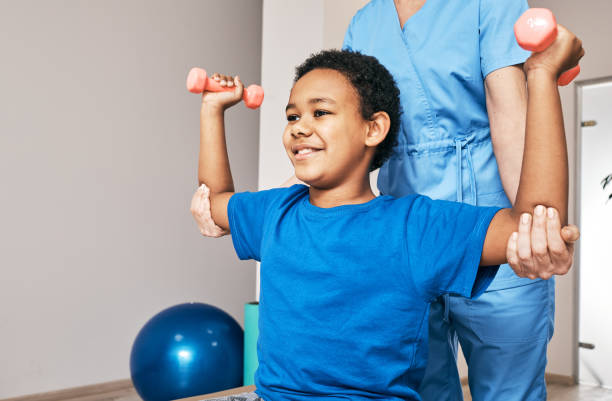 African American boy trains with physiotherapist using dumbbells at rehab center. Smiling child with hands up, using dumbbells.